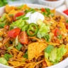 Close-up of a vibrant bowl of Doritos Taco Salad topped with sour cream, lettuce, cherry tomatoes, jalapeños, and cilantro, mixed with seasoned ground beef and crushed Nacho Cheese Doritos.