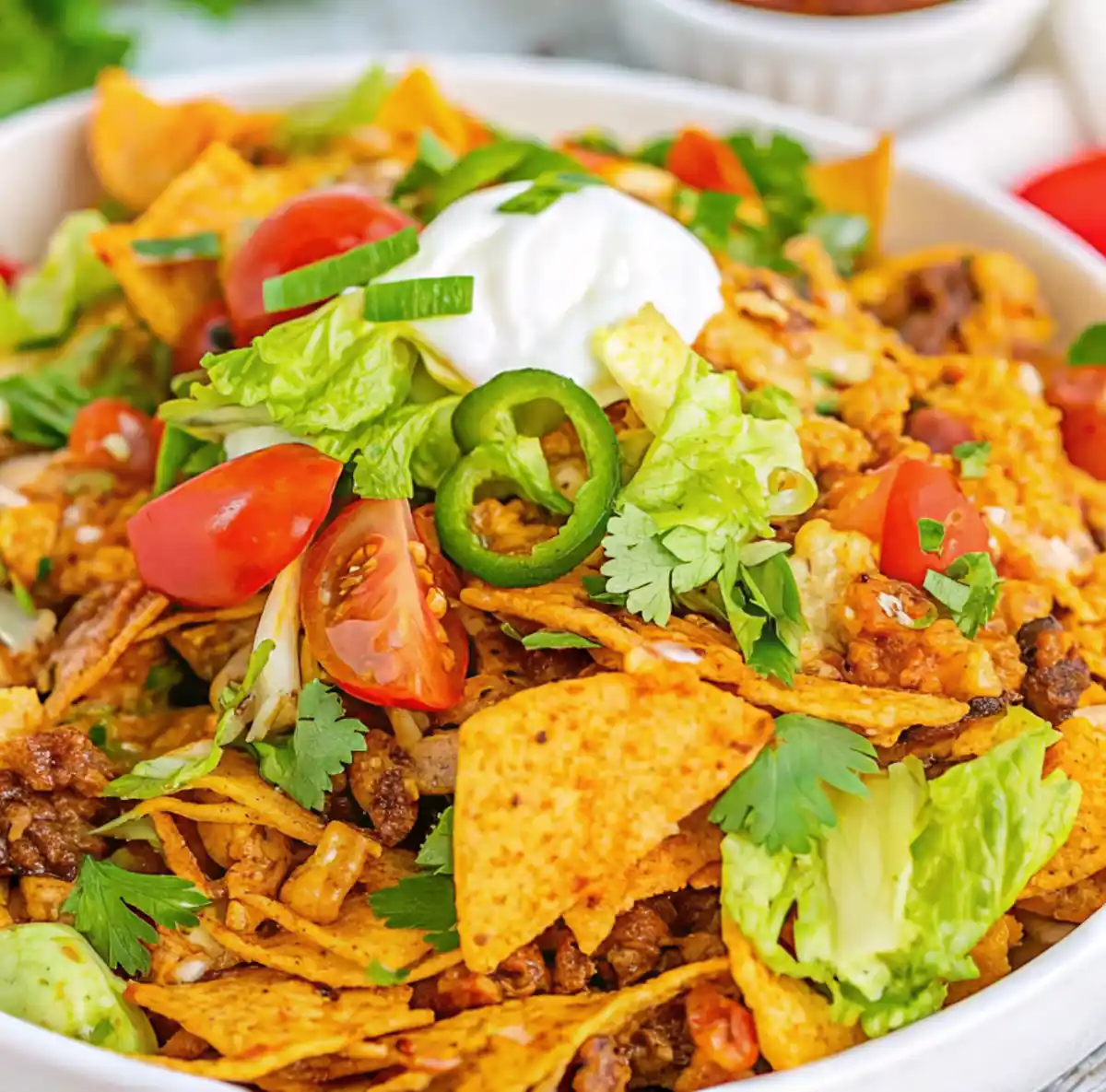 Close-up of a vibrant bowl of Doritos Taco Salad topped with sour cream, lettuce, cherry tomatoes, jalapeños, and cilantro, mixed with seasoned ground beef and crushed Nacho Cheese Doritos.