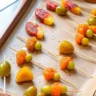 Candied fruit skewers on parchment-lined baking tray, featuring grapes, peaches, and other colorful fruit coated in sugar glaze.
