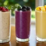 Three colorful smoothies in clear glasses on a rustic kitchen table: creamy banana oat smoothie, vibrant berry spinach smoothie, and bright tropical smoothie with pineapple and mango.