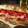 Close-up of a capicola and soppressata sandwich with provolone cheese and vinegar peppers on a seeded Italian roll, served on a wooden board.