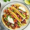 Plate of three gluten-free ground beef tacos with corn tortillas, topped with diced tomatoes, red onions, guacamole, sour cream, and fresh cilantro.