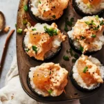 Baked sushi bites in a muffin tin, featuring rice and spicy salmon topped with creamy orange sauce, green onions, and sesame seeds.