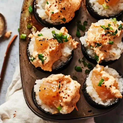 Baked sushi bites in a muffin tin, featuring rice and spicy salmon topped with creamy orange sauce, green onions, and sesame seeds.