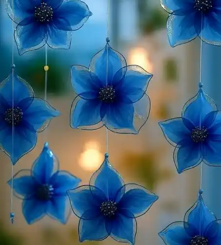 Hanging blue flower-shaped suncatchers made of translucent fabric or plastic, each with a beaded center, strung on clear lines in front of a softly lit background.