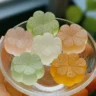 A glass bowl holding six pastel-colored, flower-shaped DIY aroma stones in shades of green, peach, white, and orange, each with detailed petal patterns.
