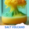 A vibrant, colorful "salt volcano" experiment in a clear glass container. Layers of oil and colored liquid create a volcano-like eruption, with a fluffy cloud of yellow and orange rising from green peaks.