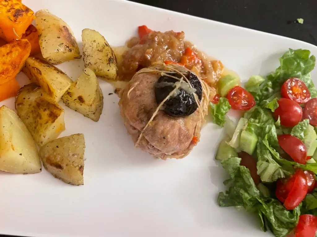 A plated meal featuring a veal paupiette topped with a prune, served with roasted potatoes, sweet potatoes, and a fresh salad of lettuce, cherry tomatoes, and cucumber.