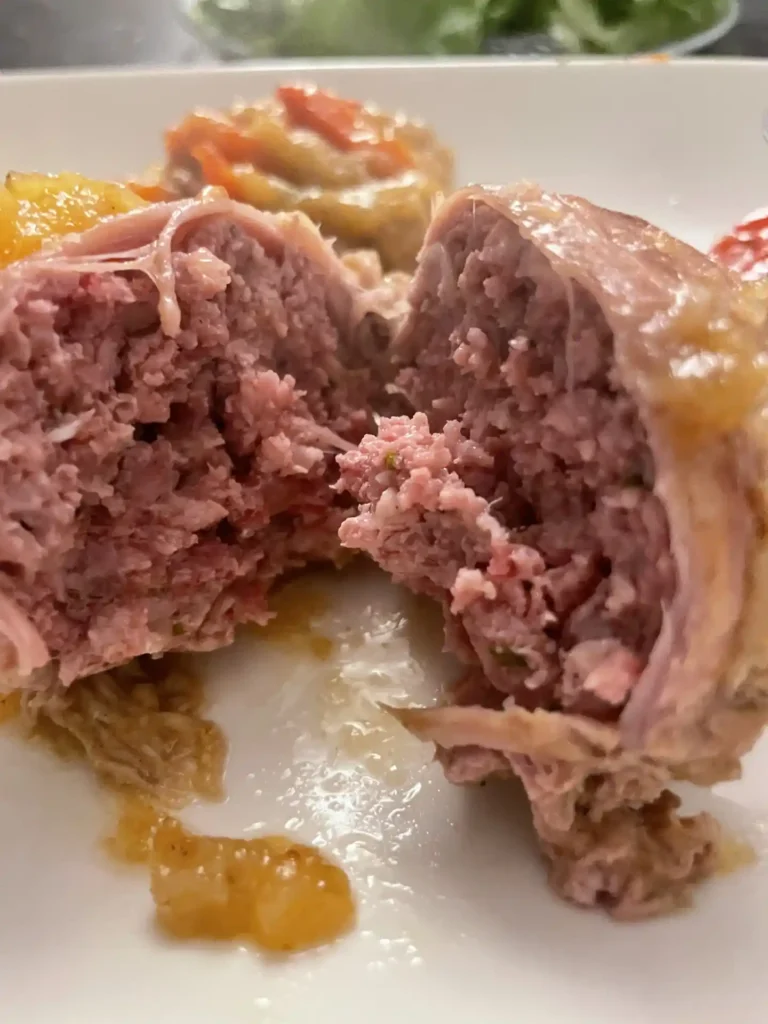 Veal Paupiettes: A Tender French Classic with a Sweet Surprise 2 A close-up view of a veal paupiette cut in half, revealing its juicy ground meat filling, with bits of onion and herbs, served with braised vegetables in the background.