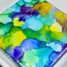 Abstract alcohol ink art on a ceramic tile with vibrant blues, greens, yellows, and purples.
